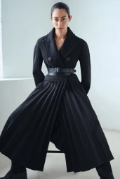 Model wearing a structured black belted coat with a pleated skirt against a plain white studio background.