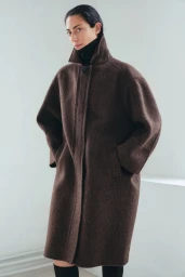 Model wearing a brown wool coat with a high collar and dark turtleneck against a plain background