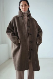 Model wearing a long brown wool coat and neutral turtleneck against a plain white studio wall