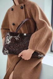 Model wearing an oversized camel wool coat and holding a brown cowhide patterned tote bag with dark leather handles.