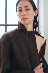 Model wearing a dark brown textured coat with a beaded brooch, photographed indoors against a window.