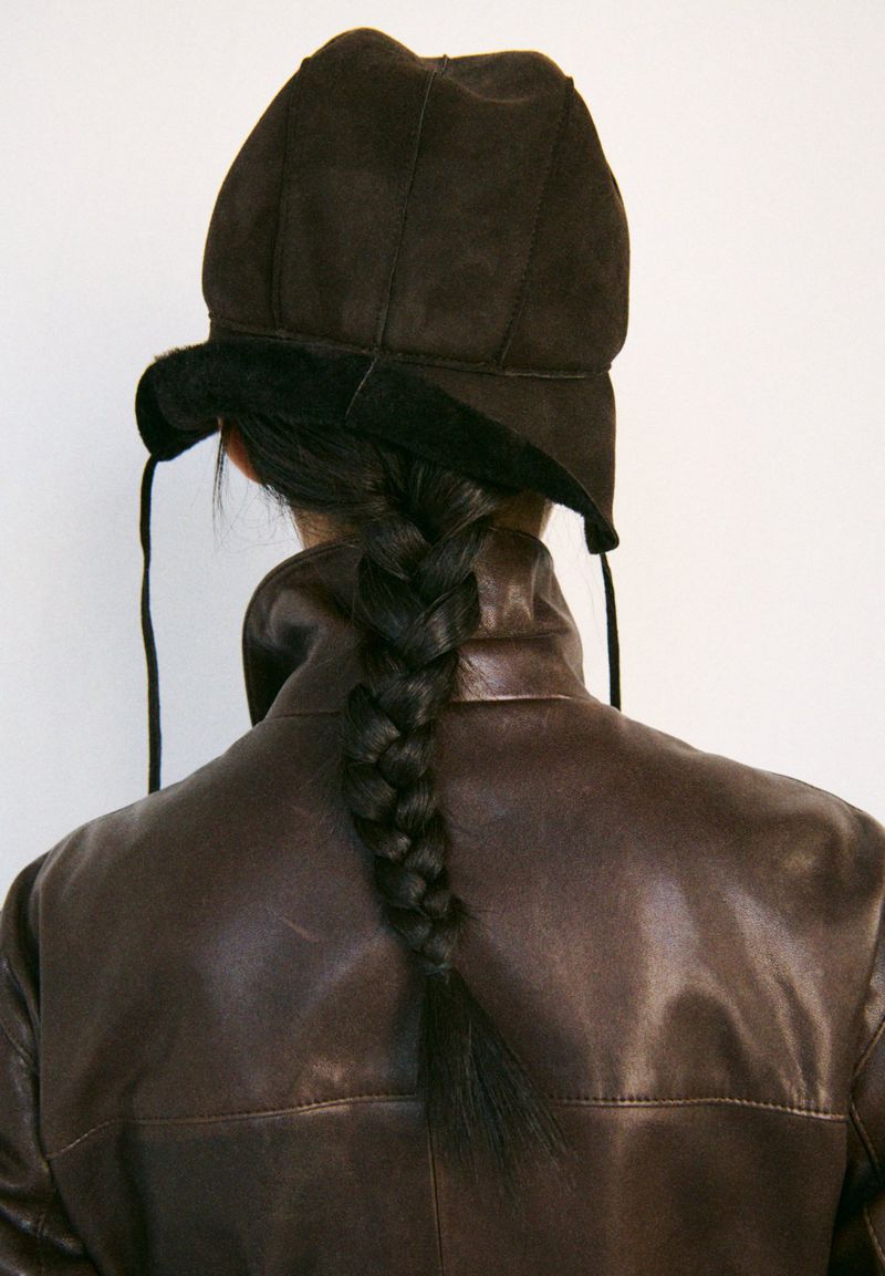 Brunette woman with a long braid wears a brown leather jacket and a dark brown hat against a plain white backdrop, studio shot.