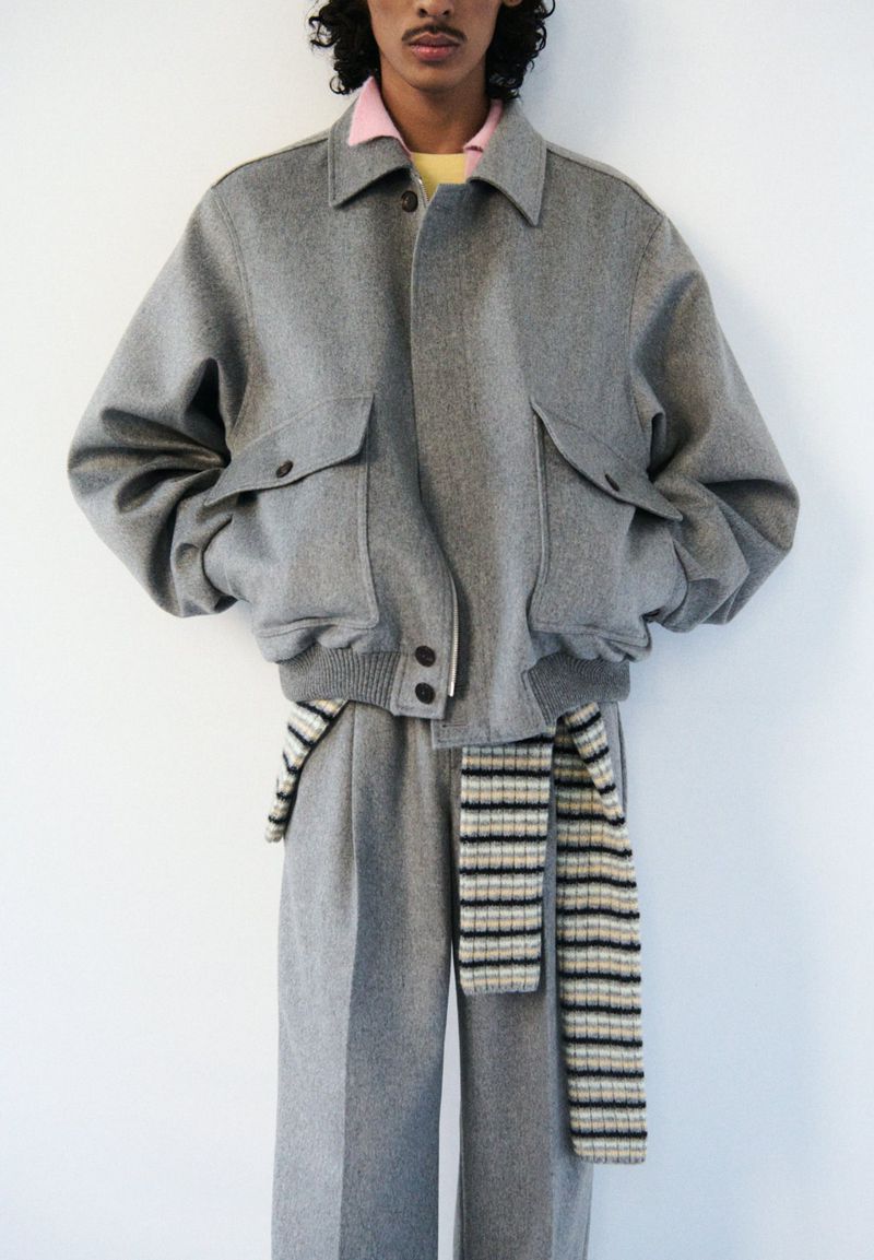 Fashion shot of a person wearing a grey wool bomber jacket with large pockets, wide-leg wool pants, and a knit scarf on a white backdrop.