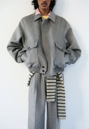 Fashion shot of a person wearing a grey wool bomber jacket with large pockets, wide-leg wool pants, and a knit scarf on a white backdrop.