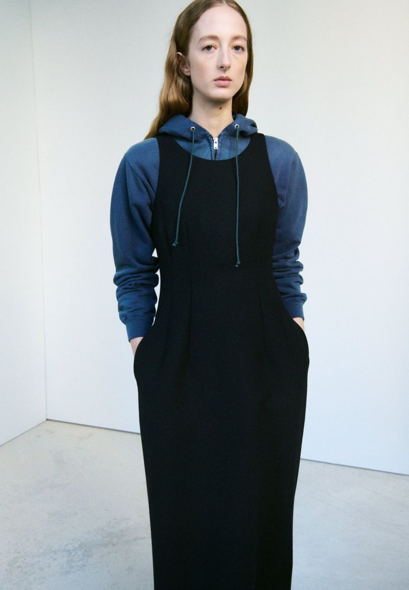 A woman in a blue hooded sweatshirt and a black maxi dress poses indoors against a neutral background in a studio shot.