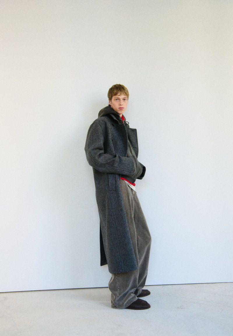 Fashionable adult man in a grey wool overcoat and loose pants, standing indoors against a white wall in soft, natural lighting.