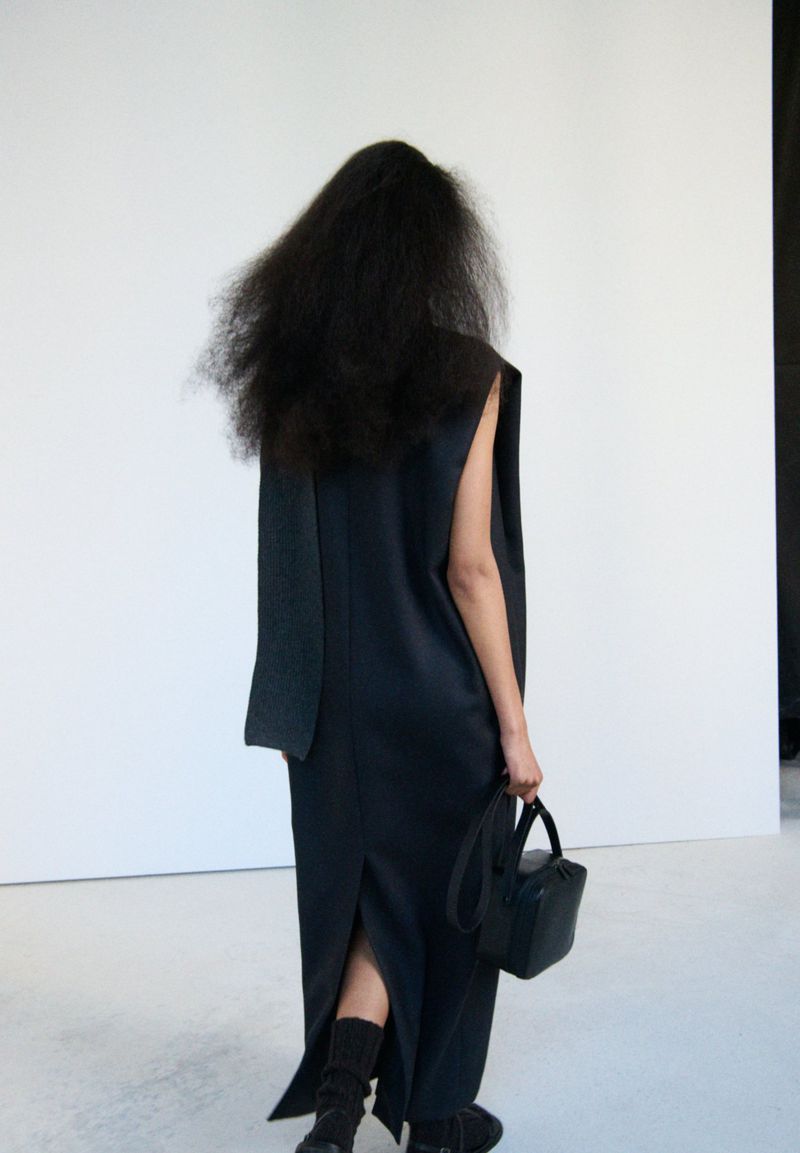 Back view of a person with dark, voluminous hair wearing a dark maxi dress and boots, holding a black handbag against a white backdrop.