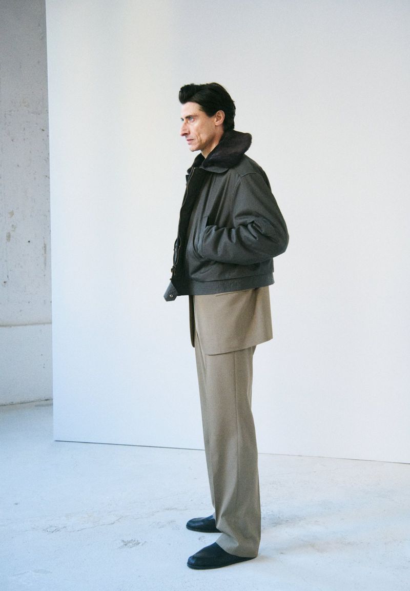 Adult man posing in a bomber jacket and khaki pants in a studio setting. The subject is facing left against a white backdrop.