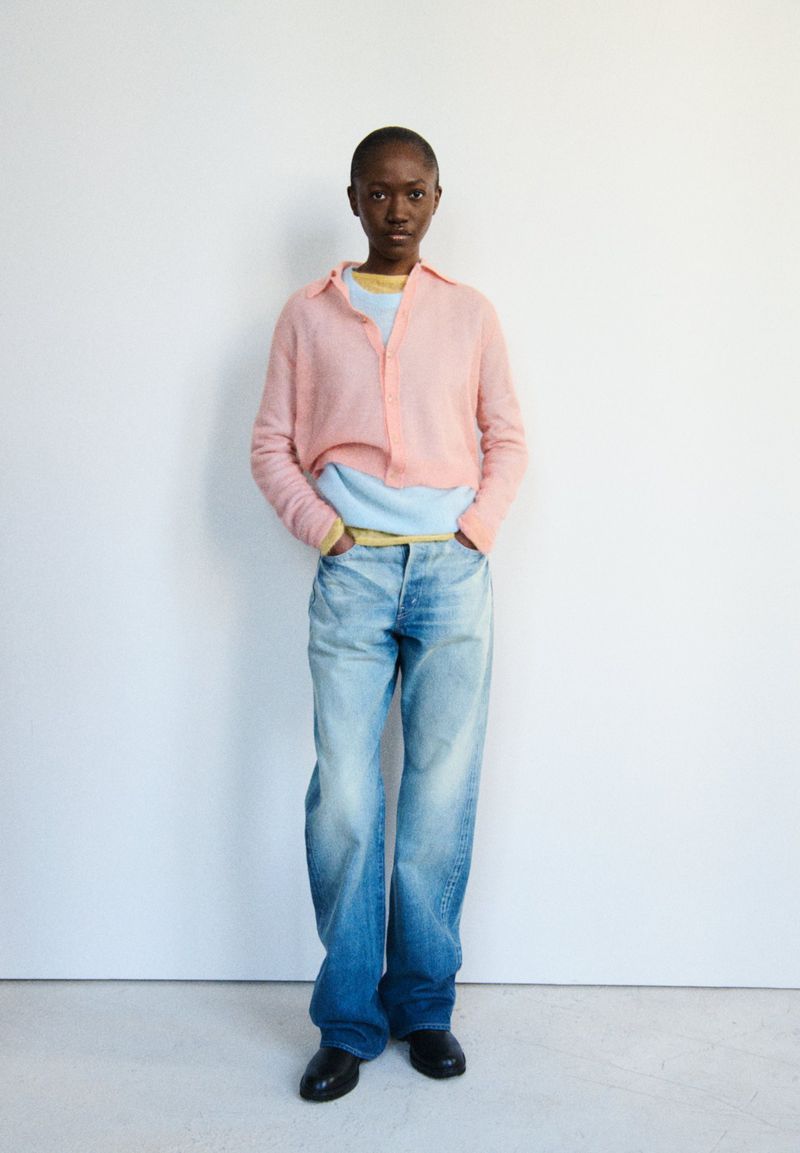 Model with short hair wearing a pink sheer knit shirt layered over a blue top and faded wide-leg jeans in a studio setting.