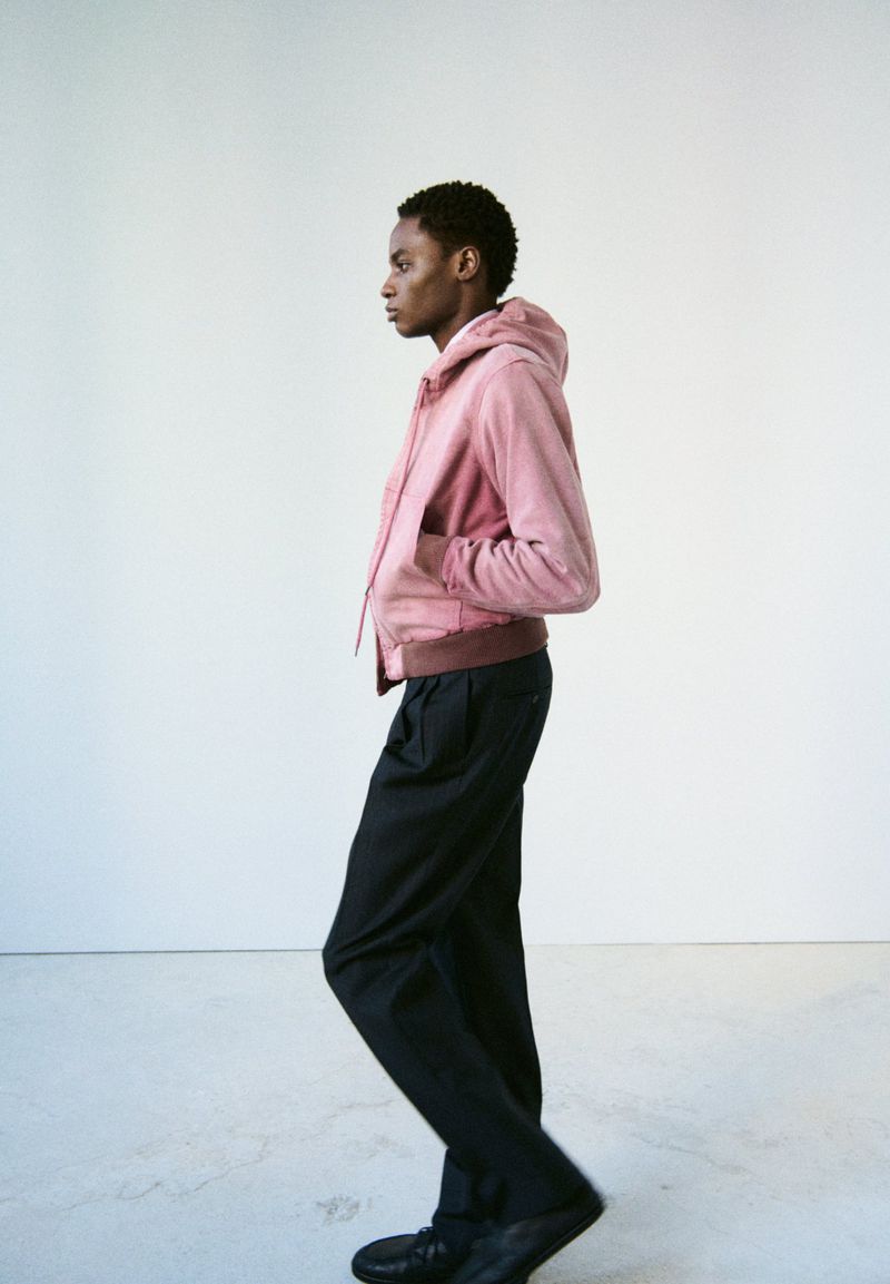Profile view of an adult Black male wearing a pink hooded jacket and black trousers walking in a white interior space, fashion shot.