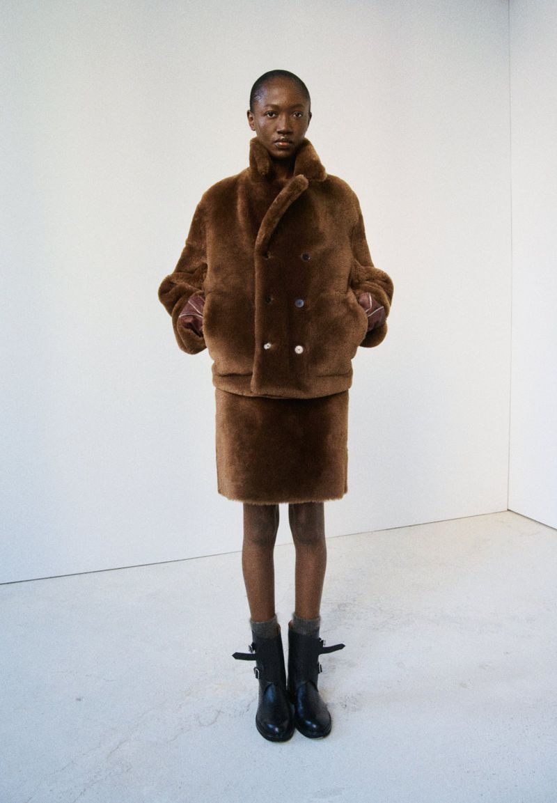A slender Black woman poses in a double-breasted brown faux fur coat and matching skirt in front of a stark white seamless backdrop.