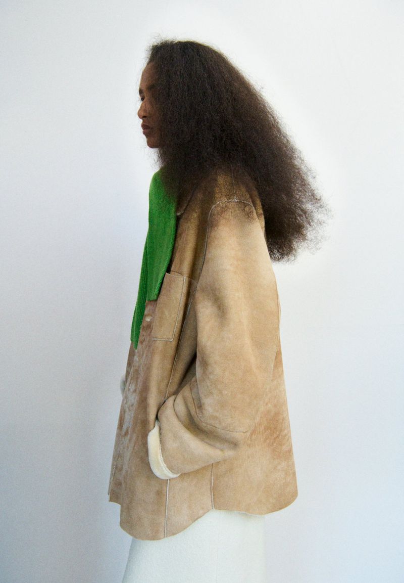 A woman with voluminous dark hair wearing a tan jacket and bright green sweater stands against a white background in a studio setting.