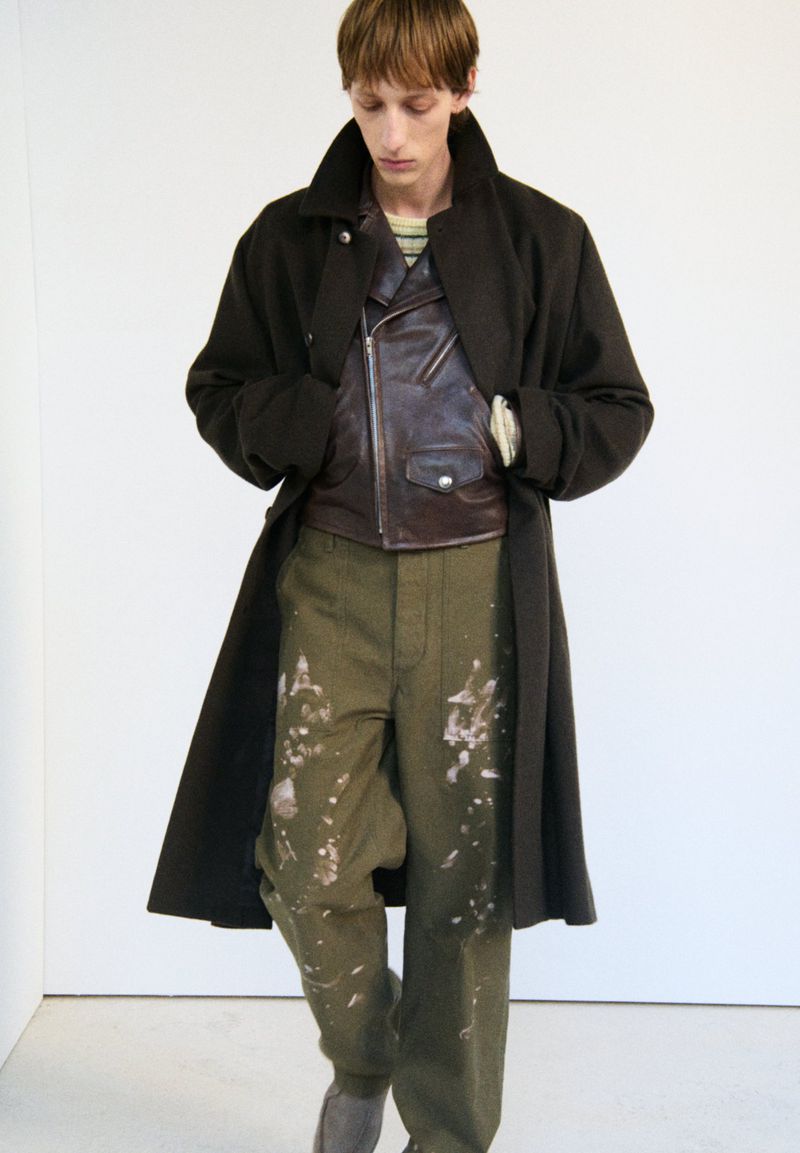 An adult male is standing indoors wearing an olive green overcoat, a brown leather jacket, and splattered pants, photographed at eye level.