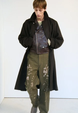 An adult male is standing indoors wearing an olive green overcoat, a brown leather jacket, and splattered pants, photographed at eye level.