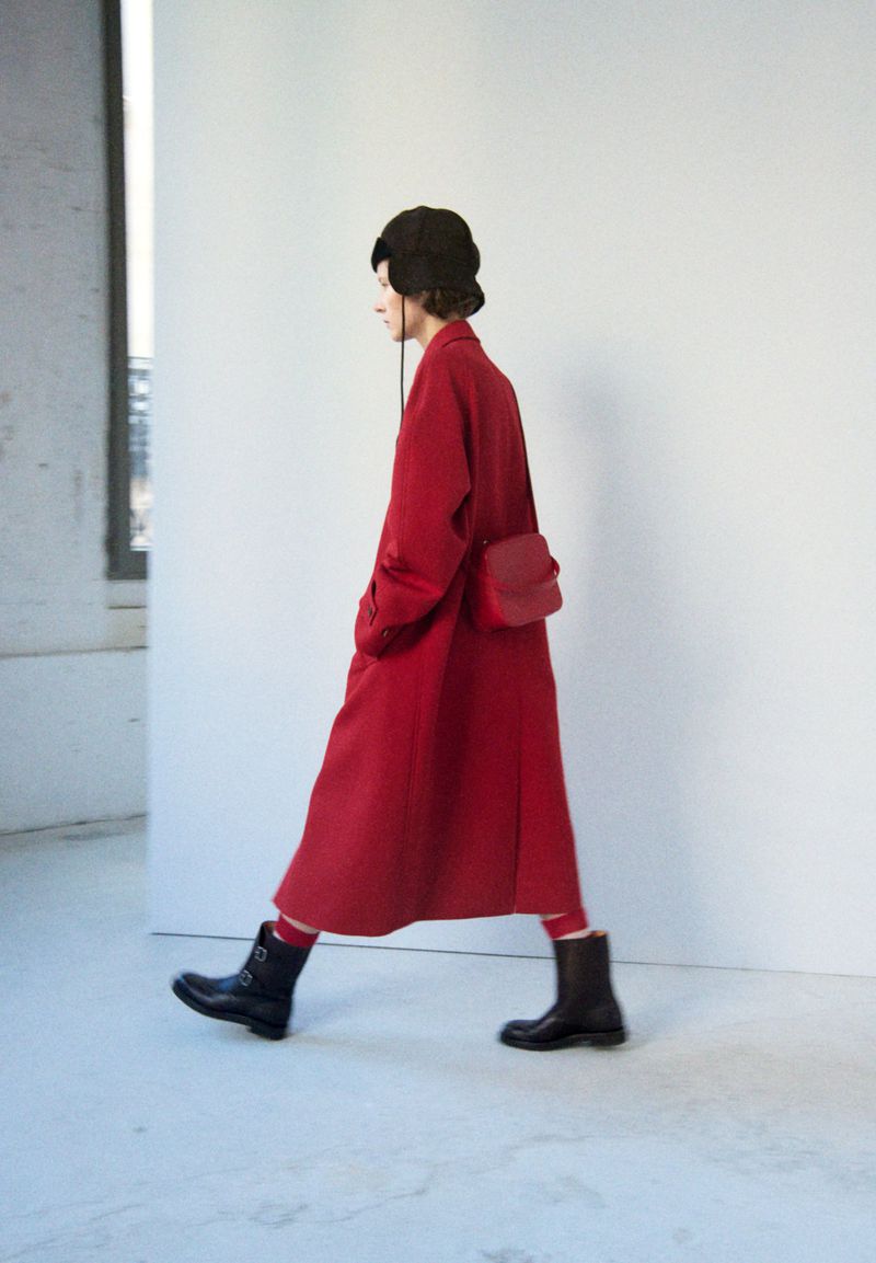 Side profile of a woman walking in a red coat with a matching bag and black boots, against a white wall with a window.