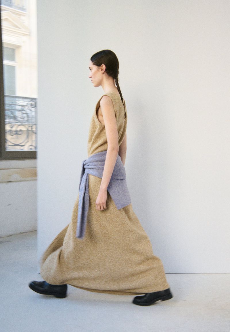 A woman in a long dress and black shoes walks past a window with a neutral background, showcasing fashion and movement.