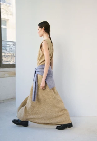 A woman in a long dress and black shoes walks past a window with a neutral background, showcasing fashion and movement.