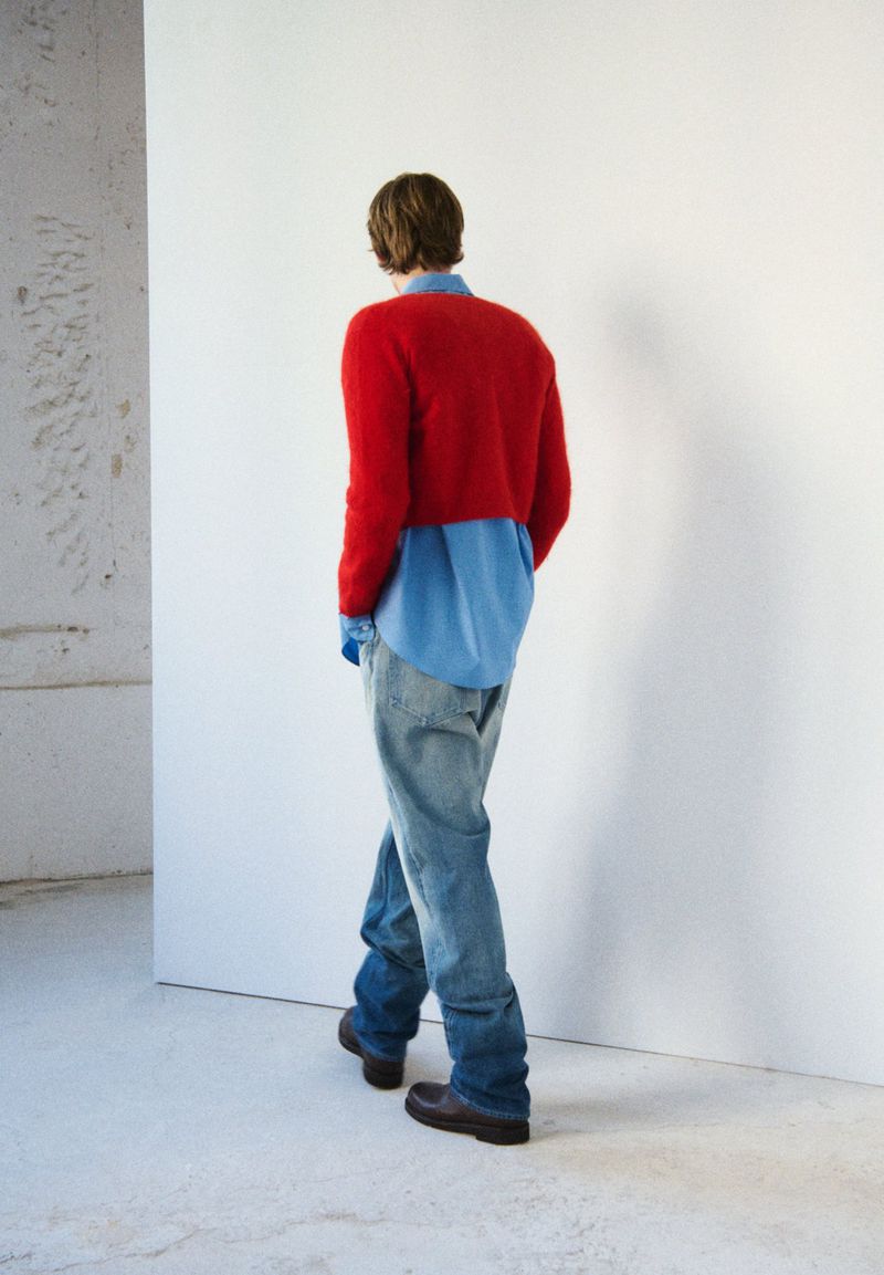 Rear view of a person in a red sweater and jeans standing in a bright white interior with concrete accents, fashion and style.