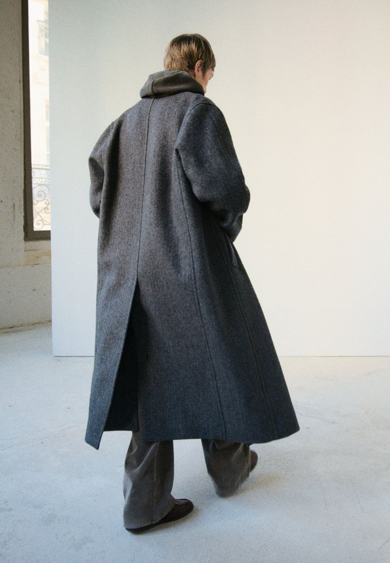 Person from the rear wearing a long gray coat with a hood, in a minimalist setting with neutral colours and natural light.