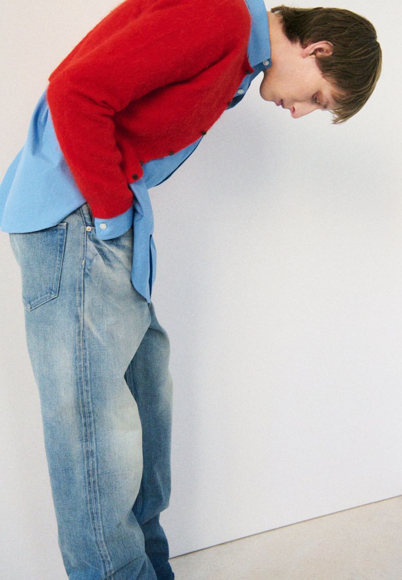 A person poses in a light blue button-down shirt, a red sweater, and blue jeans against a white backdrop in a studio setting.