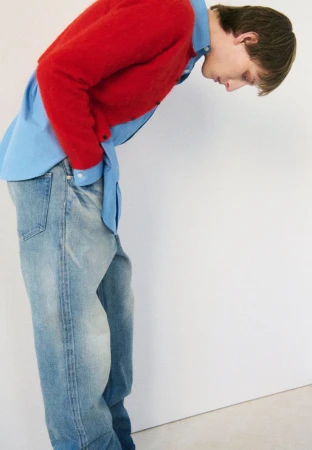 A person poses in a light blue button-down shirt, a red sweater, and blue jeans against a white backdrop in a studio setting.