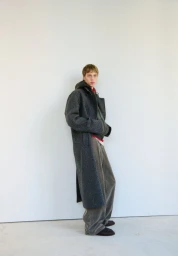 Model standing in profile wearing a long charcoal grey coat and relaxed grey trousers against a clean white backdrop.