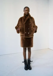 Model standing in a brown shearling jacket and skirt against a plain white studio wall and floor