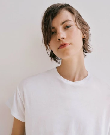 Close-up portrait of a young adult woman with short brown hair wearing a plain white t-shirt against a neutral off-white backdrop.