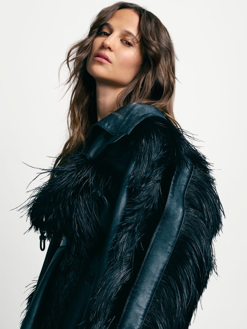 Portrait of a model with long brown hair wearing a black leather jacket with black feather detailing against a neutral background.