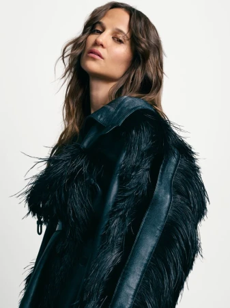 Portrait of a model with long brown hair wearing a black leather jacket with black feather detailing against a neutral background.