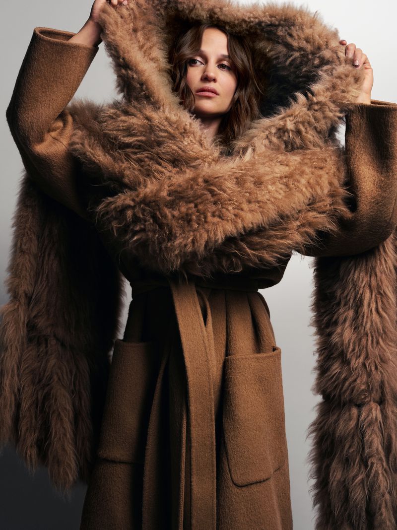 A woman models a brown wrap coat with a large fur-trimmed hood, shot in a studio with a neutral backdrop for a fashion editorial.
