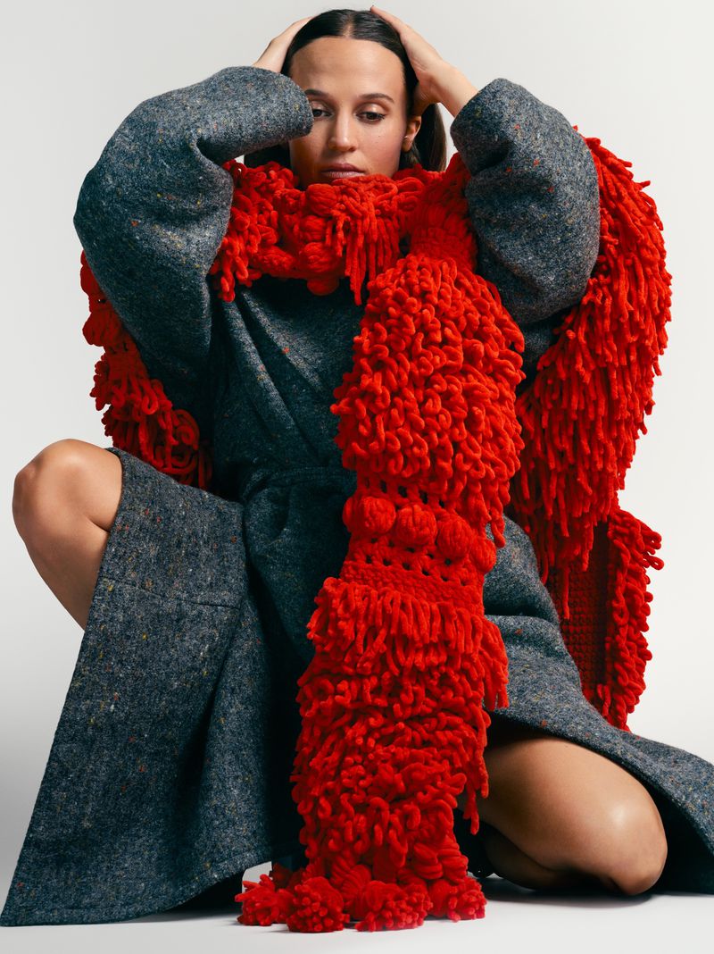 Woman in a grey coat and a large red scarf poses against a neutral backdrop, holding her head, in a studio shot.