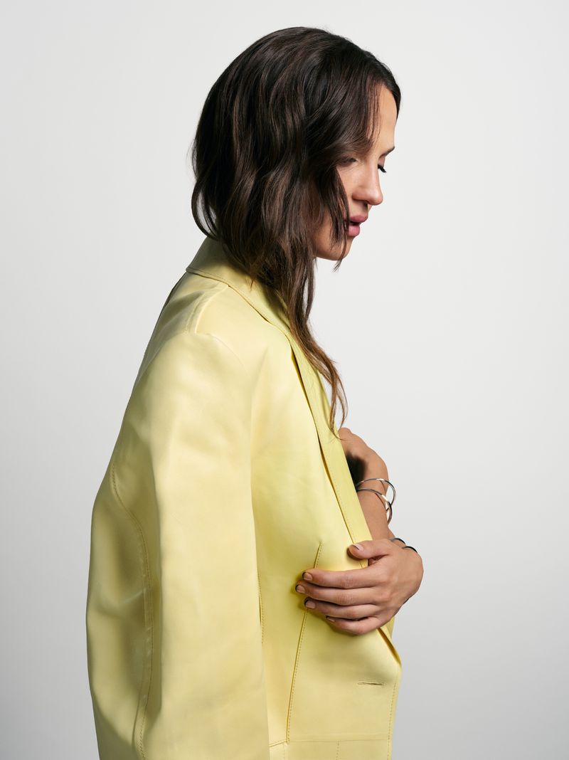 Side profile of a woman in a yellow blazer, with dark wavy hair and light skin, posing in front of a neutral background.