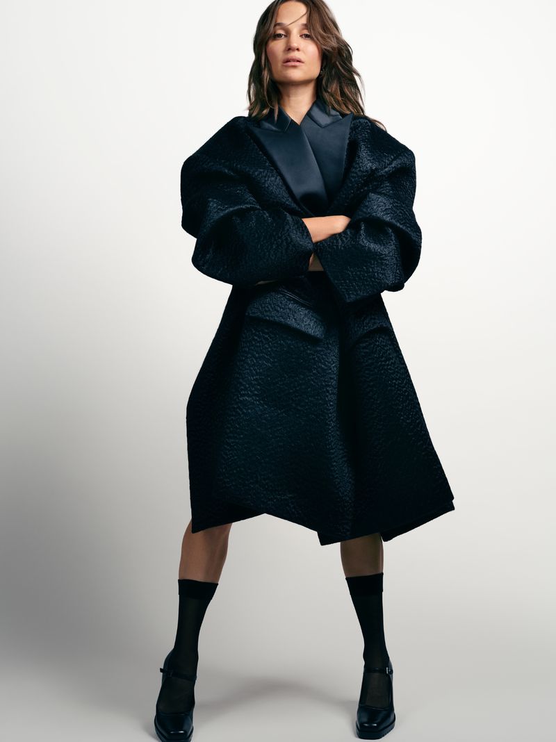 Woman in a studio portrait wearing a dark textured, oversized coat, black socks, and Mary Jane shoes against a white backdrop.