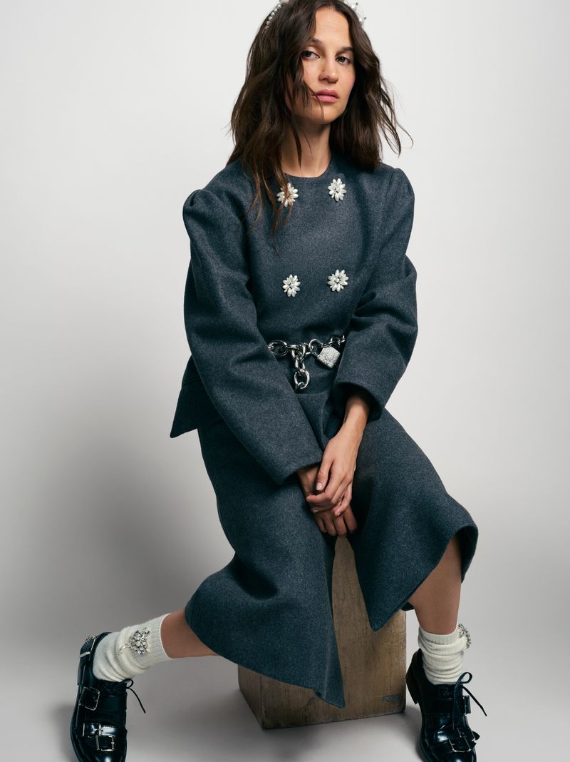 Woman in a gray coat and skirt sitting on a wooden stool, wearing black shoes and white socks, posing in front of a white wall.