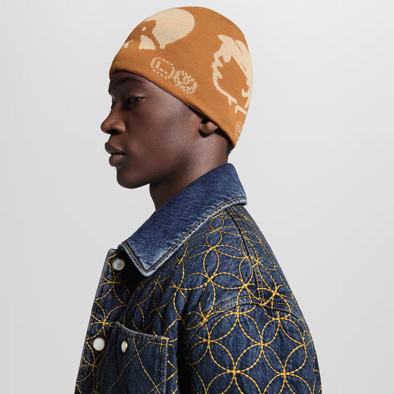 Profile shot of a dark-skinned adult male wearing a brown beanie with beige pattern, blue denim jacket with circular yellow stitching against white.