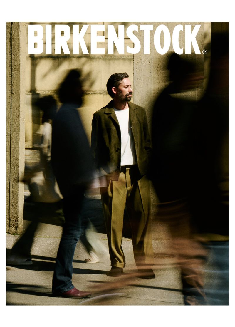 A person stands on a city street in a Birkenstock advertisement. They're wearing a blazer and tan pants, surrounded by blurred figures.