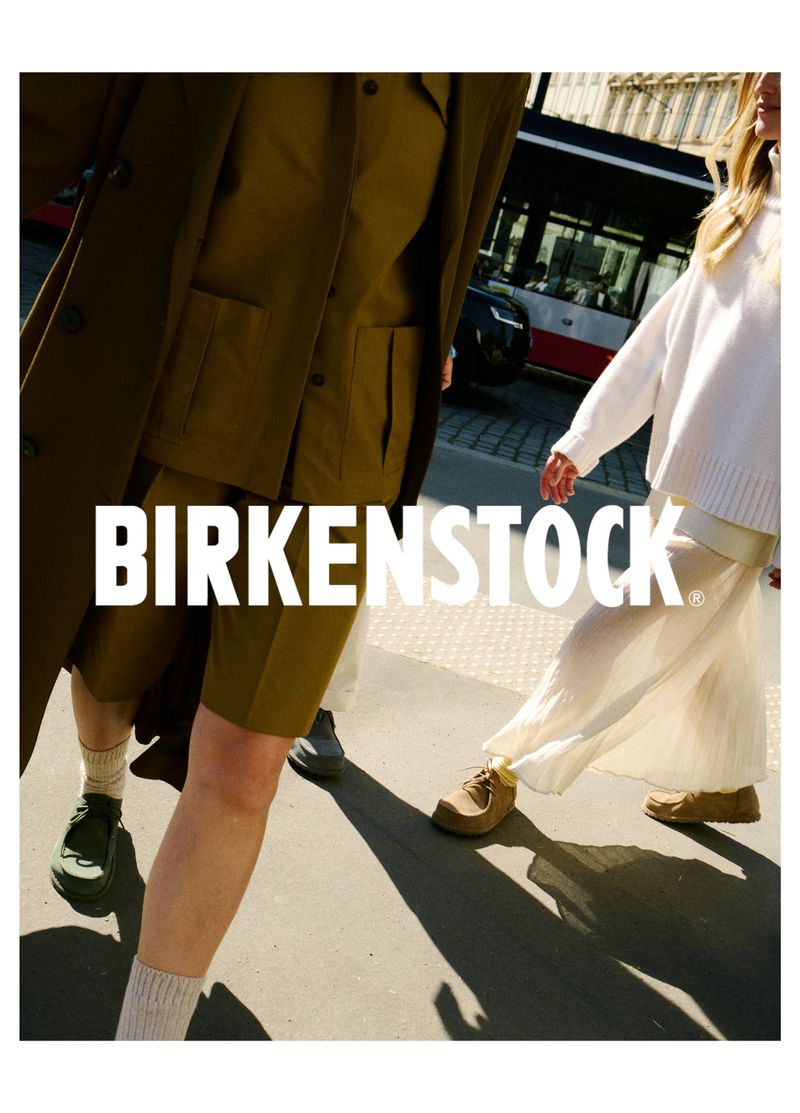 Two fashion models in olive green and white outfits are walking down a city street wearing Birkenstock shoes, advertising the brand.