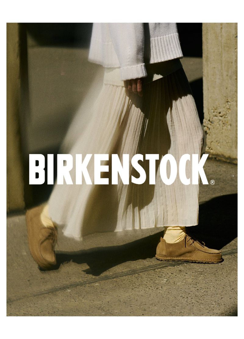 Person wearing a long white skirt, sweater, socks, and Birkenstock shoes, walking on concrete with a concrete wall in the background.