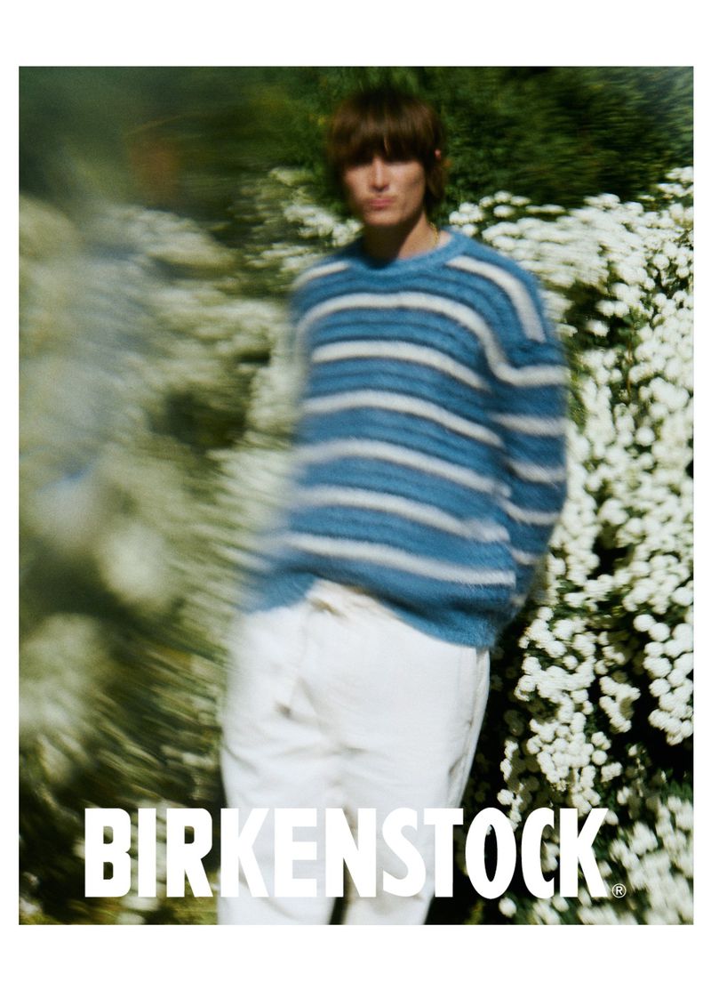 A young adult poses wearing a striped sweater and white pants in a fashion shot against a backdrop of blurred greenery and white flowers.