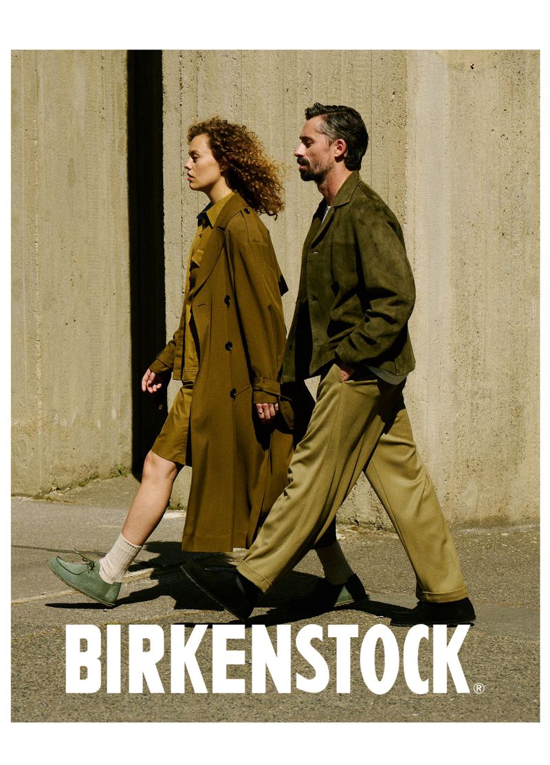 Fashionable couple walking outdoors in Birkenstock shoes. Man in olive jacket and woman in trench coat. Concrete wall background.
