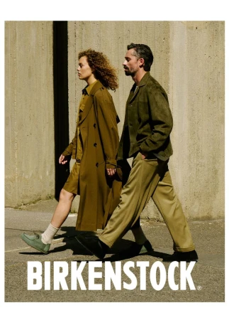 Fashionable couple walking outdoors in Birkenstock shoes. Man in olive jacket and woman in trench coat. Concrete wall background.