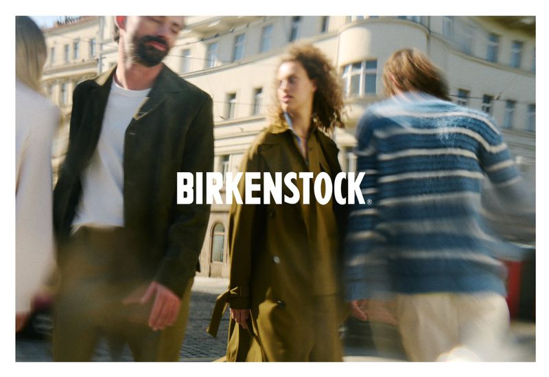 Birkenstock brand advertisement featuring three blurred adults walking across a city street wearing olive and blue outerwear.