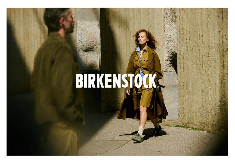 Two fashion models showcase Birkenstock footwear and apparel in a sunlit urban setting with tan columns, highlighting earthy tones and textures.