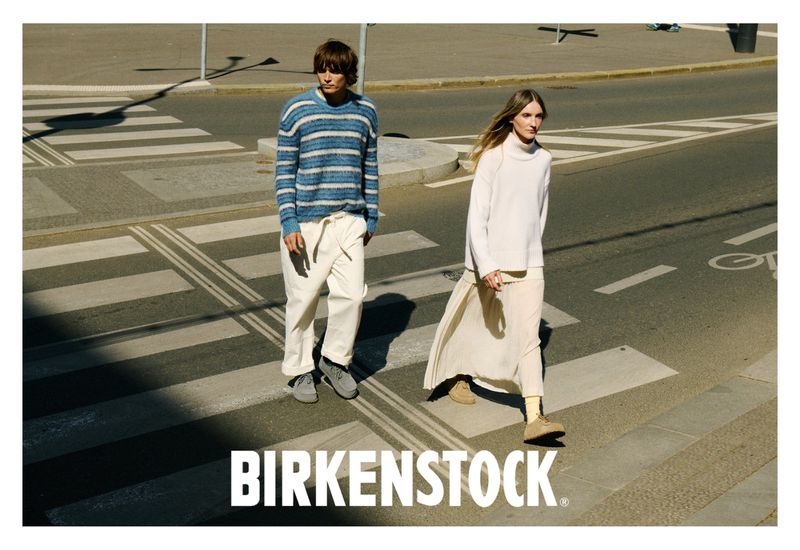 A couple in casual clothing crosses a city street on a sunny day, showcasing a Birkenstock shoes campaign with bright natural light.