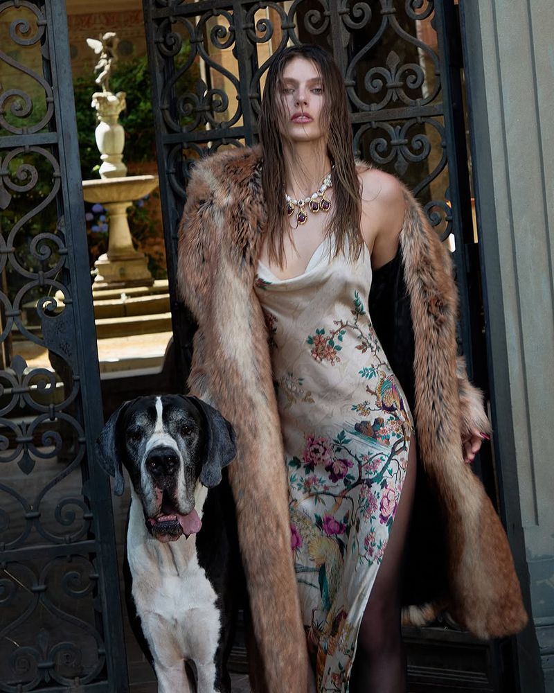 Woman in floral dress and fur coat posing with Great Dane dog near ornate iron gate and stone fountain in outdoor setting.