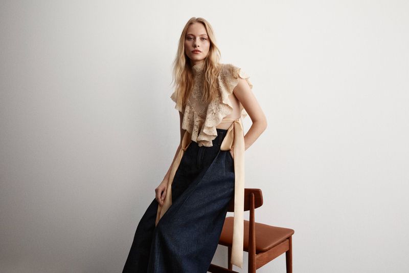 Blonde woman in a lace top and wide-leg denim pants sits on a wooden chair in a brightly lit studio with a minimalist white wall.