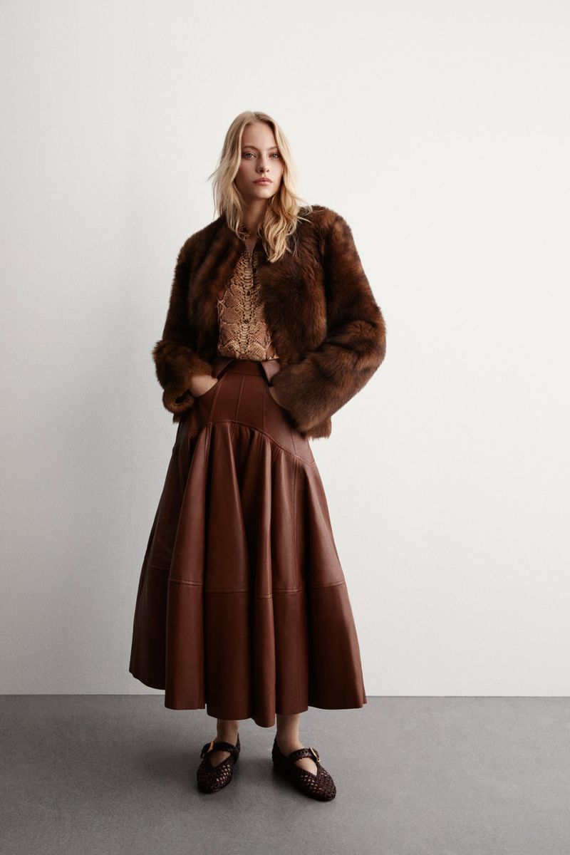 Woman wearing a brown fur coat and brown leather skirt poses in a studio setting. She also wears brown shoes and a patterned top.
