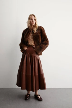Woman wearing a brown fur coat and brown leather skirt poses in a studio setting. She also wears brown shoes and a patterned top.