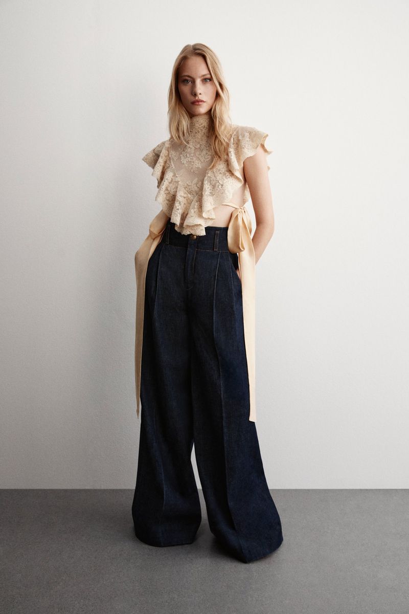 Blonde model wears a tan ruffled blouse and wide, high-waisted, dark-blue pants against a white background in a fashion studio setting.